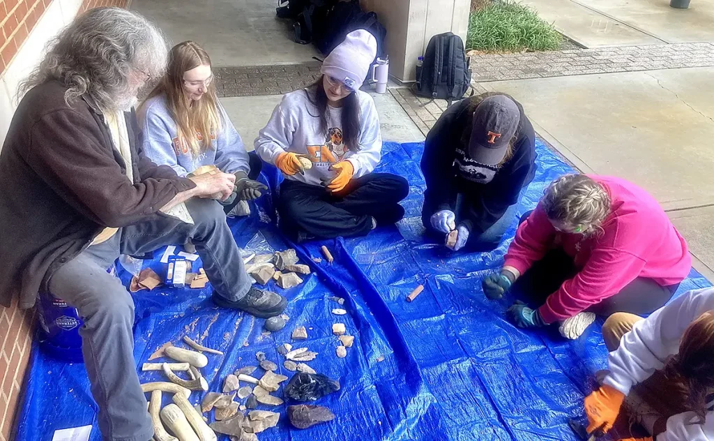 A group of students working on flintknapping while sitting on a blue tarp with bones in front of them